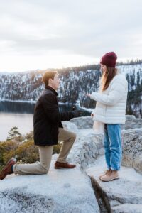 A man kneels and proposes to a woman outdoors on a rocky ledge during a Lake Tahoe Winter Proposal, with snow-covered trees and a lake in the background.