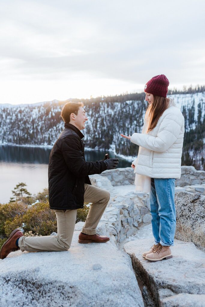 A man kneels and proposes to a woman outdoors on a rocky ledge during a Lake Tahoe Winter Proposal, with snow-covered trees and a lake in the background.