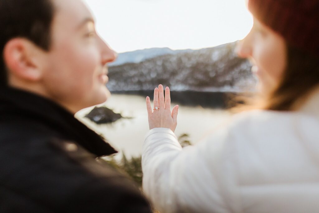 Two people outdoors, one proudly showing an engagement ring on their hand, with scenic Lake Tahoe and snow-capped mountains in the blurred background—a magical Lake Tahoe winter proposal moment.