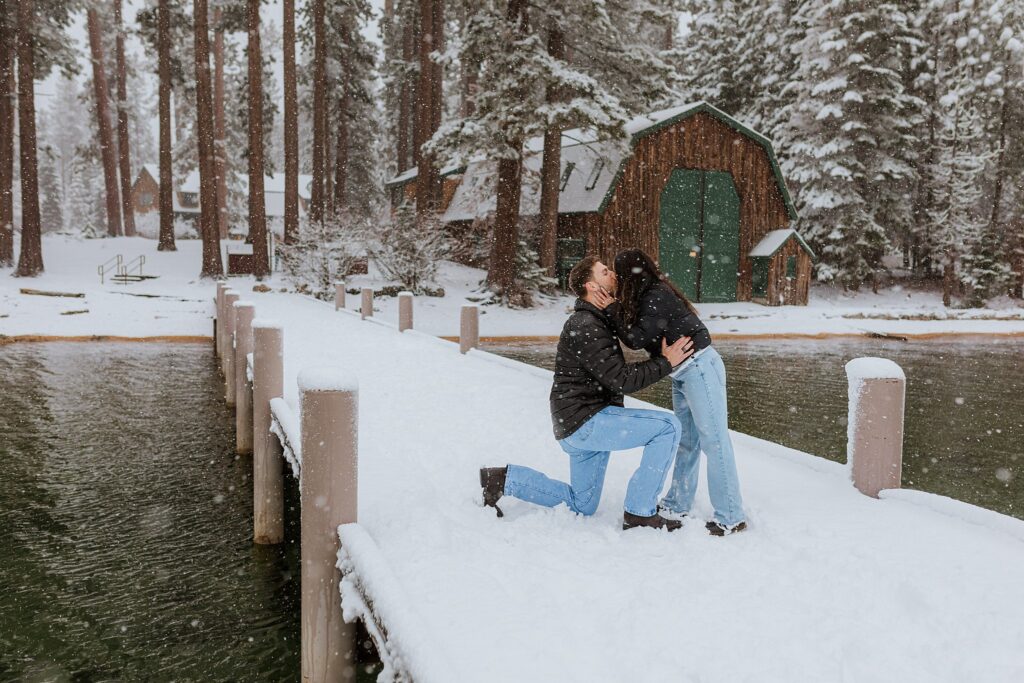 A person kneels on a snowy dock during a Lake Tahoe Winter Proposal, proposing to another who leans in for a hug, with trees and a cabin in the background.