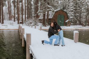 A person kneels on a snowy dock during a Lake Tahoe Winter Proposal, proposing to another who leans in for a hug, with trees and a cabin in the background.