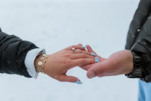 A person with light blue nails and a diamond ring on their finger holds hands with another, both in dark winter jackets, against a snowy backdrop—capturing the magic of a Lake Tahoe Winter Proposal.