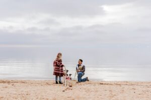 A man kneels on a sandy beach at Lake Tahoe, holding a ring box and proposing to a woman as their dog stands between them, with calm water and a cloudy sky in the background—capturing the magic of a Lake Tahoe Proposal.