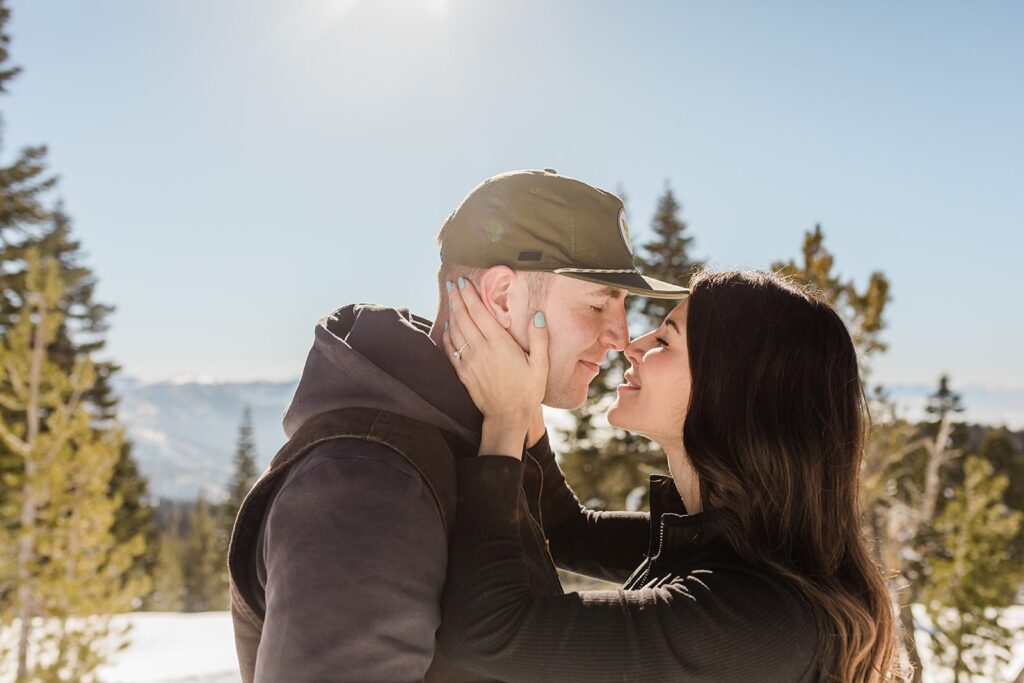 A woman and man stand close together outdoors in a snowy, mountainous area, facing each other and smiling—capturing a magical Lake Tahoe Winter Proposal with trees and blue sky in the background.