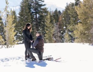 A man kneels in the snow, surrounded by pine trees and winter scenery, making a heartfelt Lake Tahoe Proposal to his beloved.