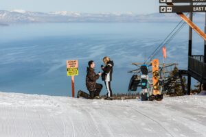 A man kneels to propose to a woman on a snowy ski slope near an “Expert Skiers Only” sign, with snowboards and a ski lift in the background—capturing the thrill of a Lake Tahoe Winter Proposal.