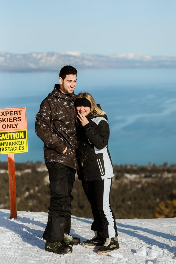 Two people in winter clothing stand on a snowy slope by a sign reading "Expert Skiers Only," overlooking a stunning lake and mountains—capturing the thrill of an unforgettable Lake Tahoe Winter Proposal.