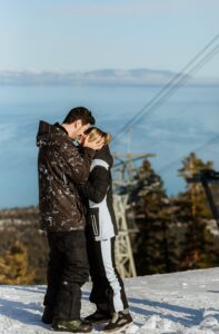 A couple in winter clothing stands close together on a snowy mountain with trees, a ski lift, and a lake in the background, capturing the magic of a Lake Tahoe Winter Proposal.