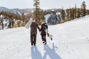 Two people dressed in winter gear walk hand in hand on a snowy slope, each carrying a snowboard, with trees and mountains in the background—capturing the magic of a Lake Tahoe Winter Proposal.