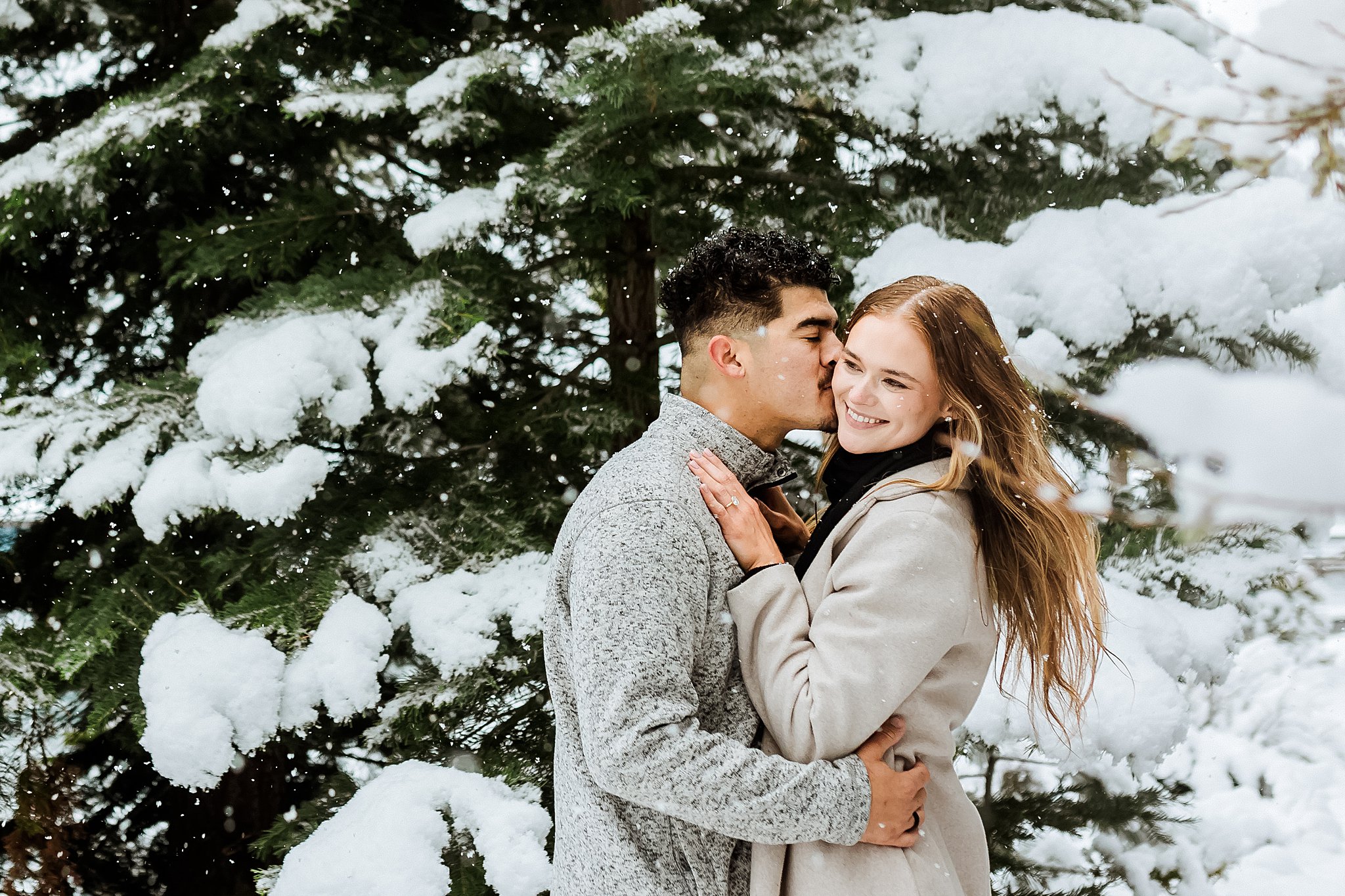 A man kisses a woman on the forehead as they embrace in front of snow-covered evergreens during a romantic Lake Tahoe Winter Proposal.