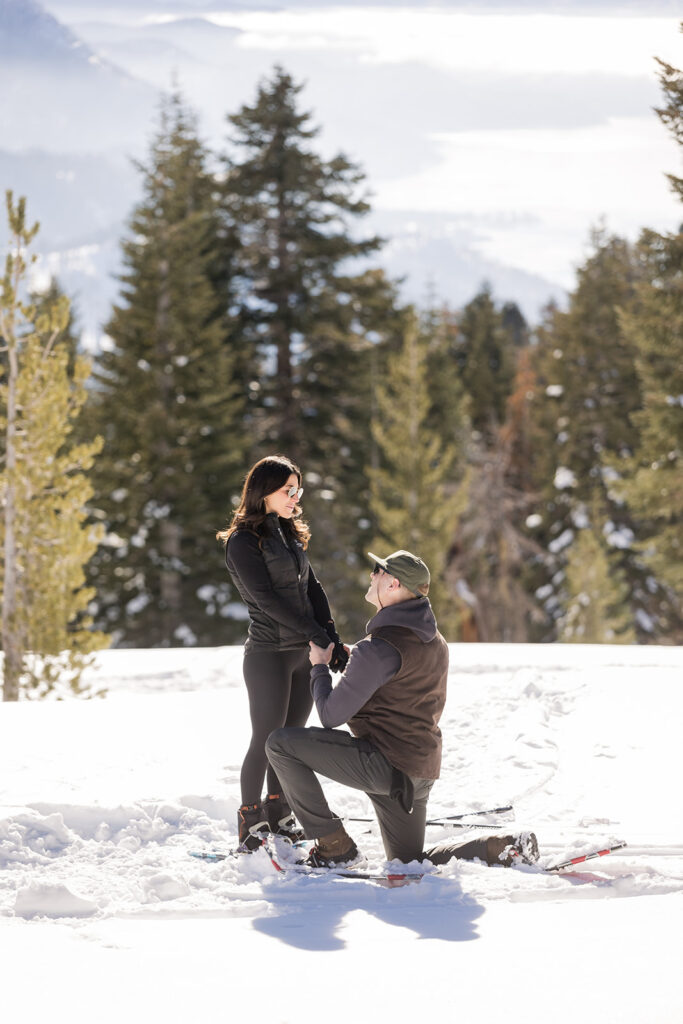 A man kneels in the snow proposing to a woman, with snowy trees and mountains in the background—a magical Lake Tahoe Winter Proposal.