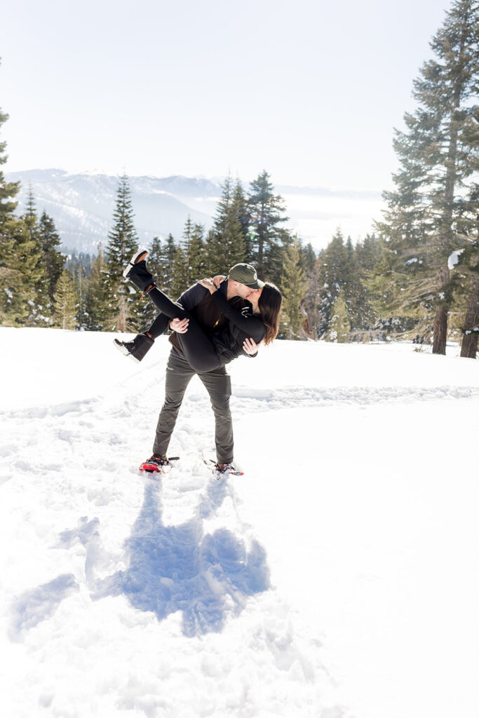 A person lifts and kisses another in a snowy landscape surrounded by pine trees, with mountains visible in the background, capturing a magical Lake Tahoe Winter Proposal.