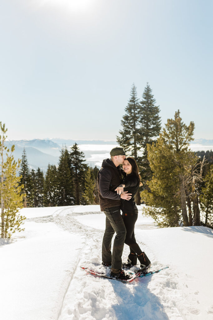 A couple stands close together on snowshoes in a snowy mountain landscape near evergreen trees beneath a clear sky, capturing the magic of a Lake Tahoe Winter Proposal.